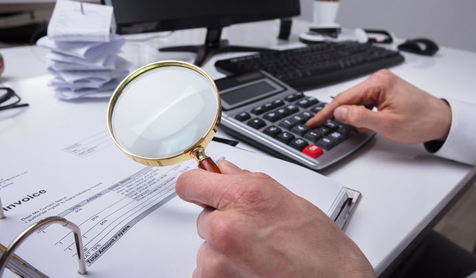Close-up Of A Businessperson's Hand Examining Invoice Through Magnifying Glass On Desk